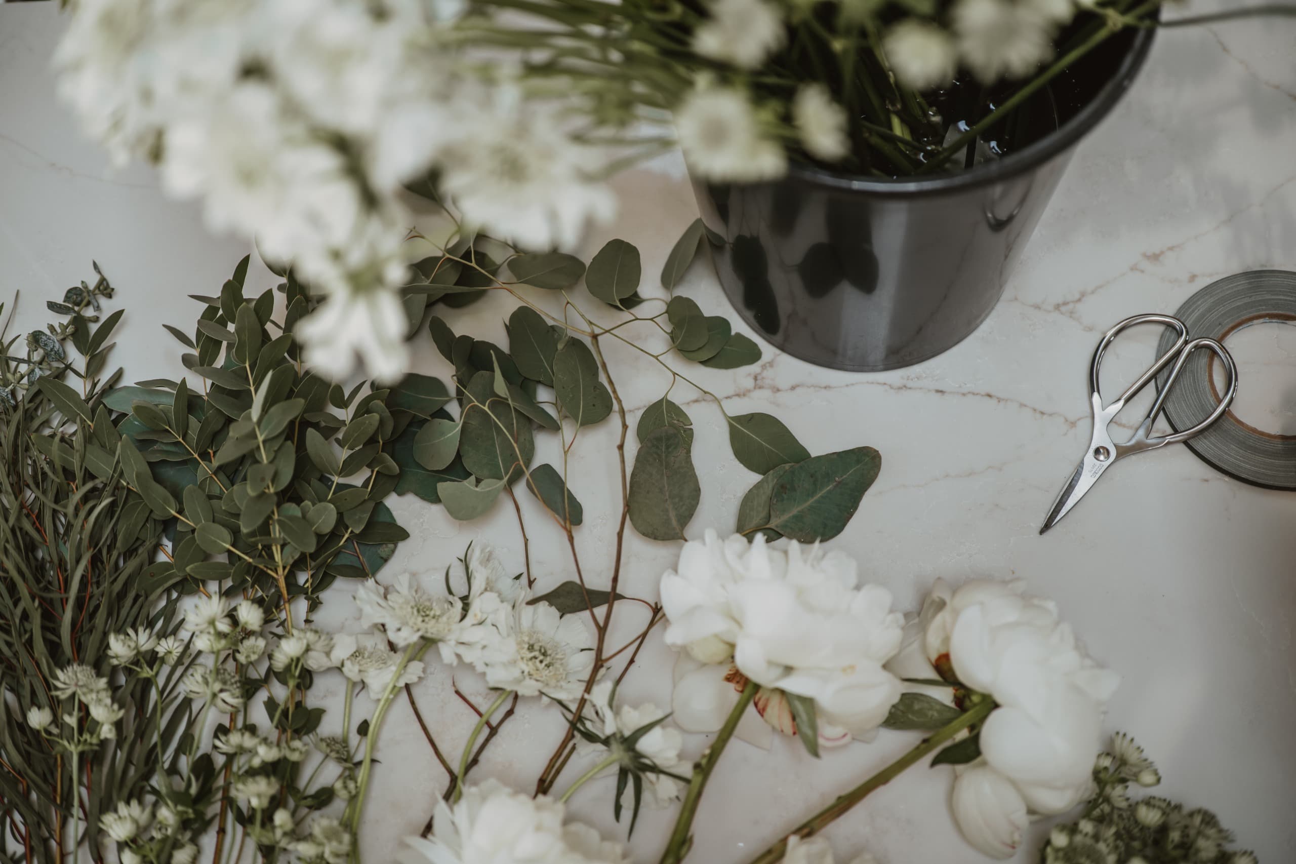 Eucalyptus stems, white flowers and a dark florist's bucket on a marble surface, beside scissors and a reel of tape.