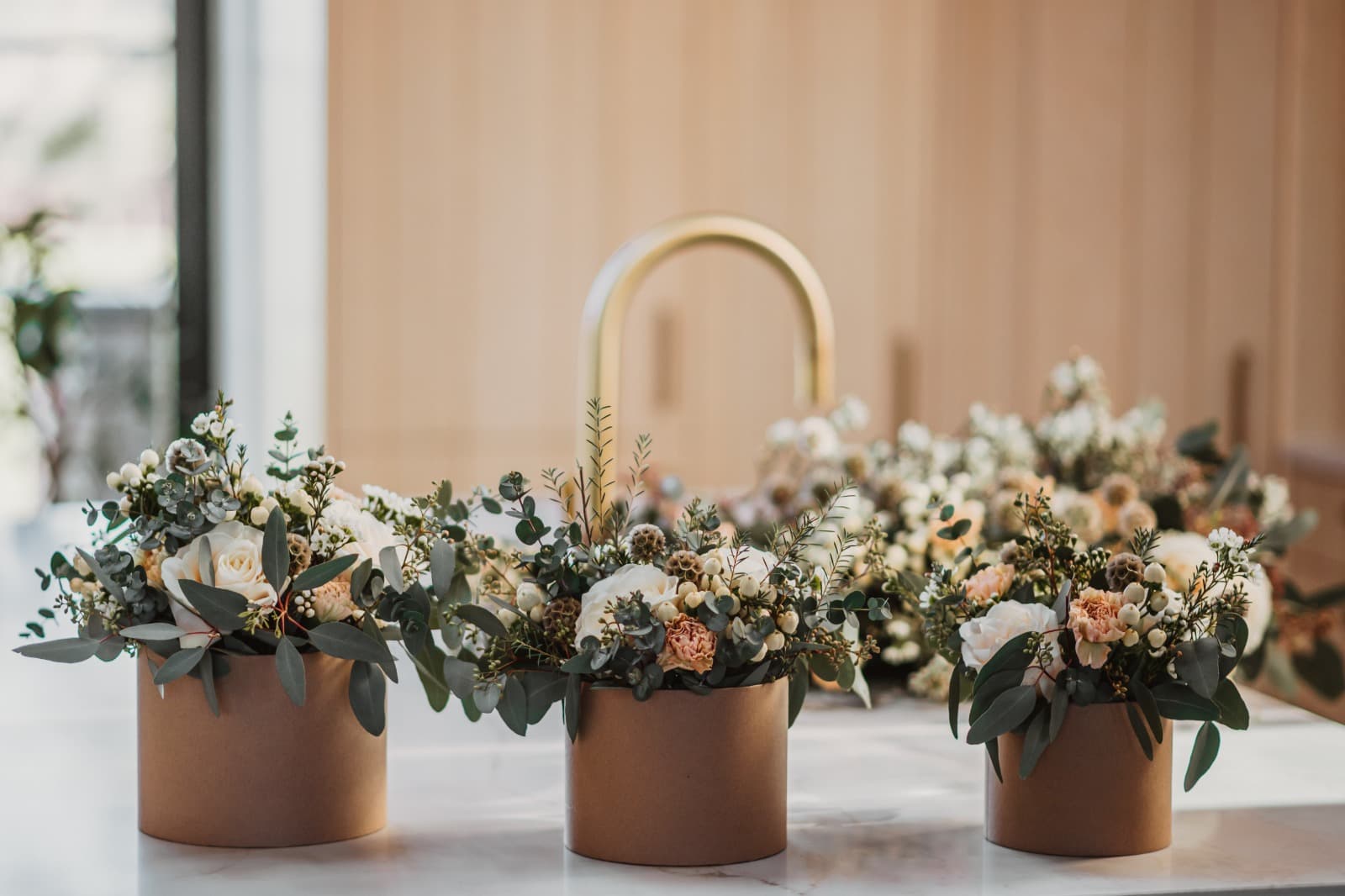 Three kraft-paper wrapped flower arrangements of white, cream and peach blooms with eucalyptus, standing in a row on a marble counter in front of a curved brass tap.